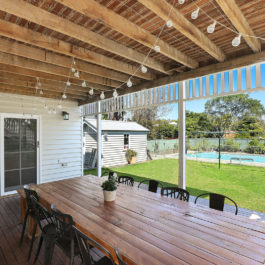 Une jolie terrasse en bois prend place sous un auvent. Une grande table rectanguaire en bois est entourée de chaises de jardin en métal. Au loin on voit une piscine entourée d'une pelouse verte.