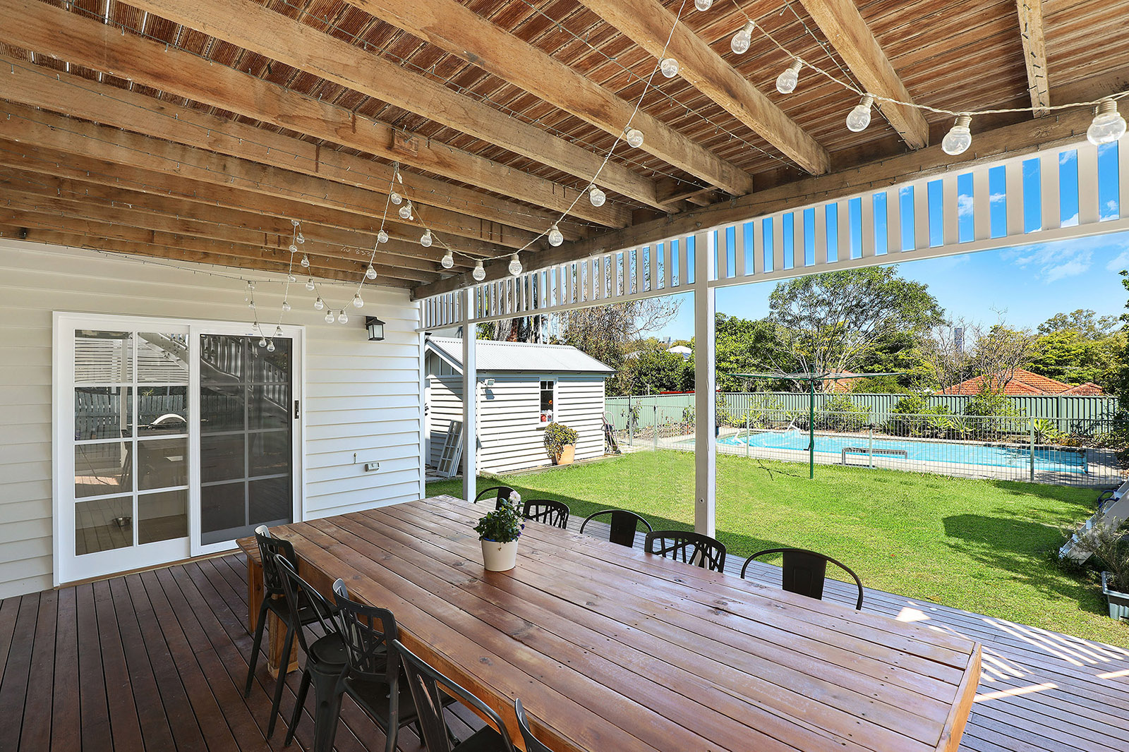 Une jolie terrasse en bois prend place sous un auvent. Une grande table rectanguaire en bois est entourée de chaises de jardin en métal. Au loin on voit une piscine entourée d'une pelouse verte.